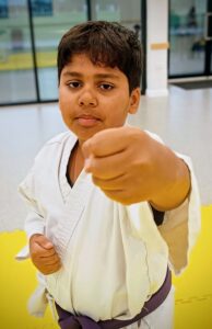 A close up of a young boy in karate unform with purple belt looking at the camera and punching towards the camera
