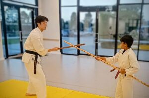 A karate instructor and a young student cross bo staff on a yellow matt