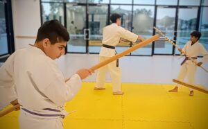 A young man in a karate uniform holds a bo staff while other karate students train with bo in the background