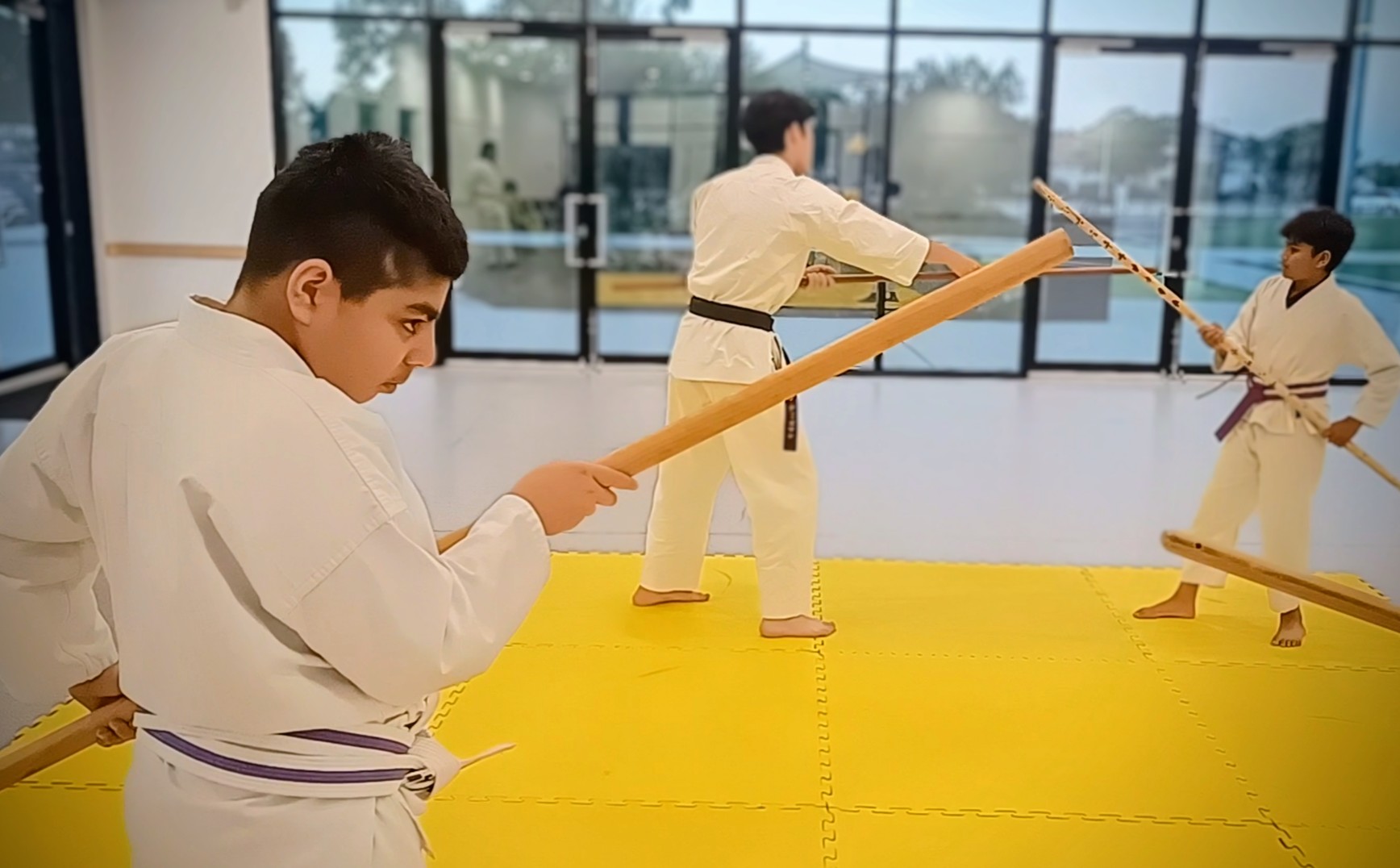 A young man in a karate uniform holds a bo staff while other karate students train with bo in the background